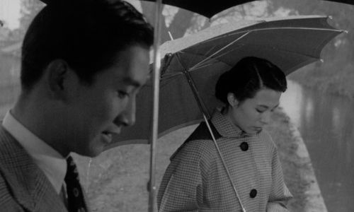 A man and woman hold umbrellas while looking at the river