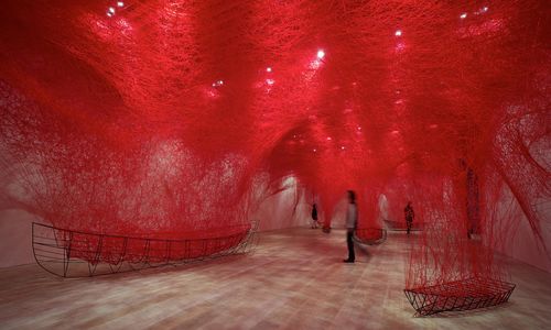 A person walking under an installation of fine red thread suspended from the ceiling and connect to small boat frames