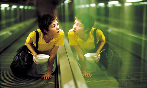 A woman kneels on a moving walkway, holding a bowl of noodles in one hand