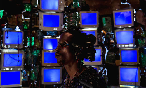 A black man seen in profile, framed by stacks of video monitors glowing blue