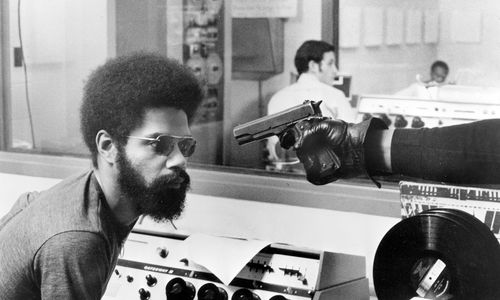 A gloved hand points a handgun at a Black man with a beard and afro