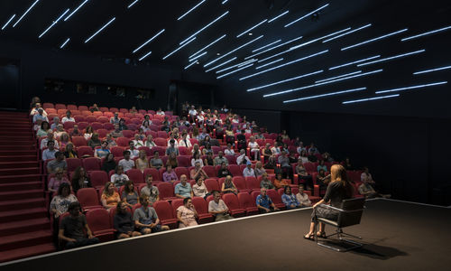 An audience sitting in a darkened theater, watching a woman on stage