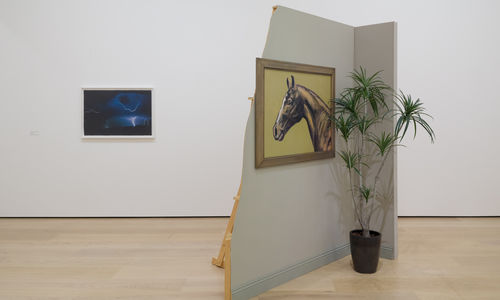 installation image with framed work on wall and a sculpture comprised of a false wall with painting and potted plant