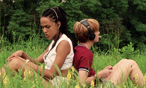 Two girls sit back-to-back in a field, wearing headphones