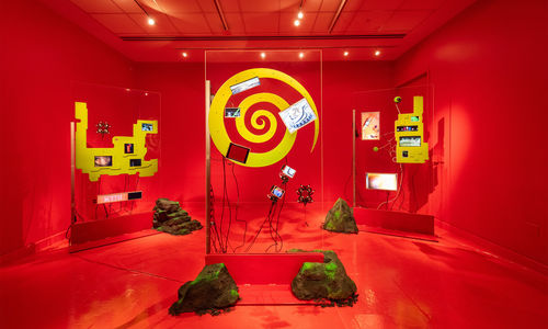 Installation image of a red room with sculptures made of transparent glass, yellow shapes, and electronic components