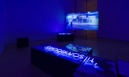 Installation with a video screen in the background showing a street scene, and a neon sign reading "til' somebody" laying on a plinth in the foreground