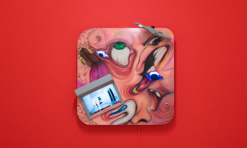 Square multi-media work with distorted images of faces and video screens, on a bright red wall