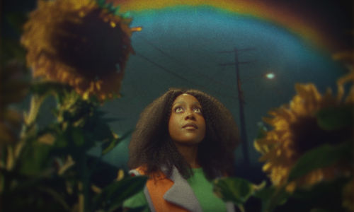 A Black woman, framed by sunflowers, looks up at a rainbow in the night sky