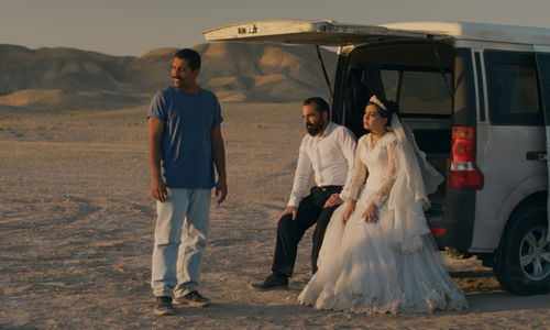 A couple dressed for their wedding sits in the open hatch of a van in the dessert