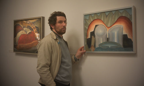 A man in a tan jacket fingers a framed painting