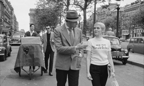A man and a suit and a woman in a New York Herald Tribune shirt walk down a city street, followed by a cameraman