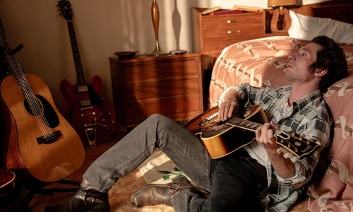 Jeffrey Allen White as Bruce Springsteen sits against a bed playing an acoustic guitar