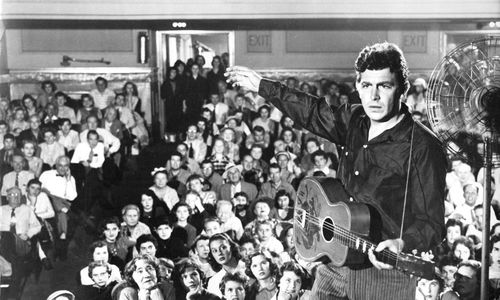 A man holding a guitar on stage, his back to a full auditorium