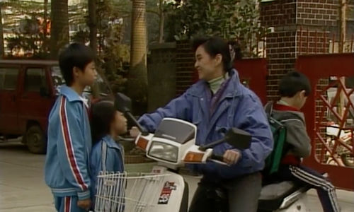 A woman on a motor scooter talks to two children in matching tracksuits