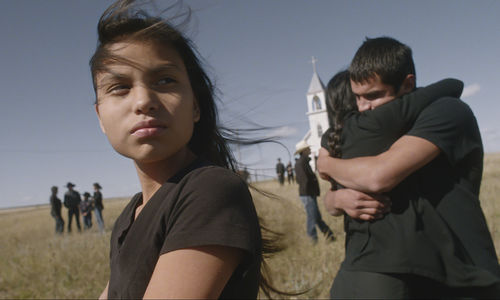 A girl looks off-camera, while behind her a man and woman hug in front of a church