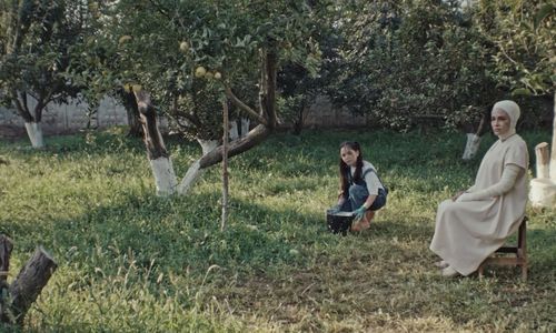 A girl kneels next to a woman in all white amidst a garden