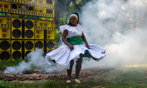 A woman in a white dress dances in front of a wall of yellow speakers, surrounded by smoke