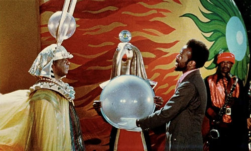 Sun Ra, in an elaborate headdress, stands with a man holding a large orb in his white-gloved hands