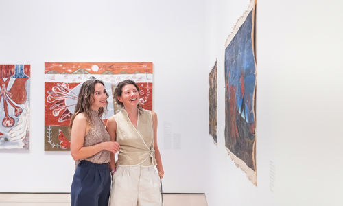 Two women look at a painting on a gallery wall together