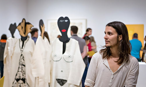 A person with long, straight brown hair and a short beard glances to the right. Just behind him are visible four mannequins wearing caftans with designs painted onto them