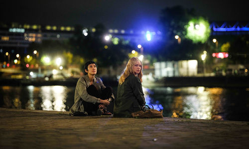 Two women sit on the waterfront, with city lights twinkling behind them