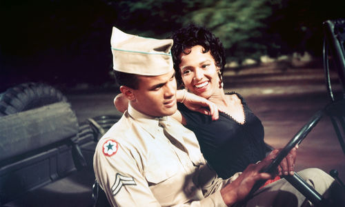 A woman in a black dress and a man in a solider's uniform sit in the front seat of a jeep, the man holding the steering wheel while the woman rests her elbow on his shoulder and smiles