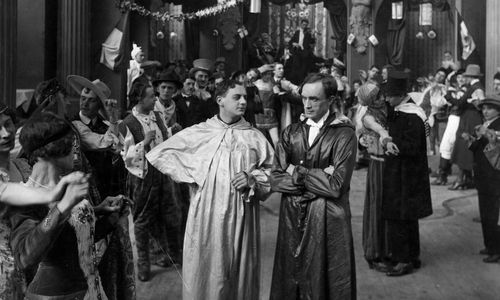 Two men in silken robes stand in the middle of a hall filled with dancing couples