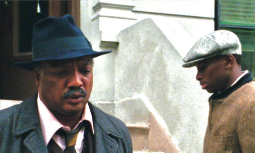 Two men, one facing the camera wearing a fedora and suit, the other facing down