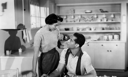 A woman stands over a man sitting at a table, pencil in hand, in a kitchen