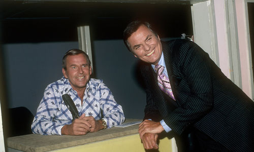 A man in a pin-striped suit smiles as he leans against a desk, where another smiling man sits in front of a microphone