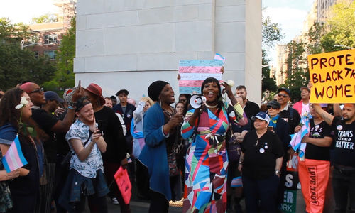 A dark skinned person with long black hair and a colorful dress speaks into a loudspeaker at an outdoor protest