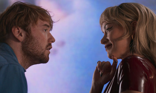 On the left, a man with short, scruffy blond hair and beard gazes into the eyes of a woman with wavy, blond hair