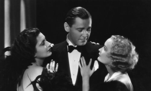 A dapper man in a tuxedo looks at a blond woman clinging to his left arm, while a dark haired woman clings to his right