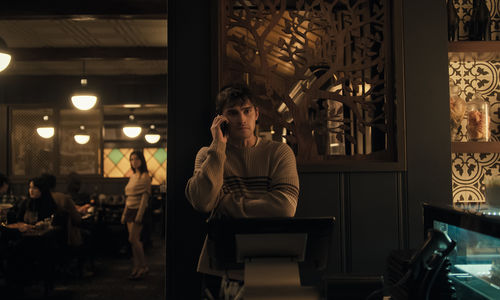 A young man in a striped sweater holds a cell phone next to his head, while out-of-focus in the background, a long-haired figure in a beige sweater and dark skirt looks his way