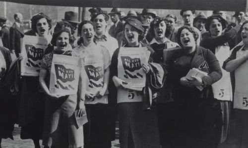 Black and white photo shows a crowd of women standing, facing the camera, with their mouths open as if shouting. Many women in the foreground hold papers against their chests that have a graphic of a flag with "All Out May 1" written on the flag.