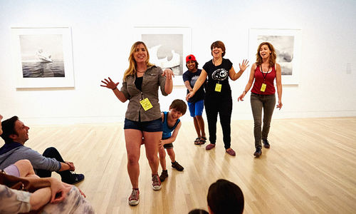 Children and adults in an art gallery, smiling and interacting. What appear to be black and white photographs are hanging on the wall.