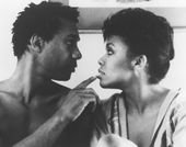 Still from the film The Brother From Another Planet (1984) showing a close-up of a shirtless man looking into the eyes of a woman in a robe and touching her lip