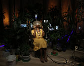 A person sits in a beauty salon chair in a dark room with potted palm trees behind her. The person has medium-dark skin, wears a yellow dress with several metal necklaces, is barefoot, and holds the chair's drying helmet over their face with two hands. Various-sized video monitors peek out rom the plants. 