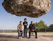 Film still from "Official Competition" (2022), showing a gigantic rock suspended over three people who seem not to notice