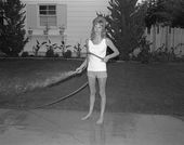 Black and white photo of a woman spraying the sidewalk with a hose
