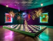 View of a gallery lit in green and pink, with a large, vibrant sculpture hanging from the ceiling