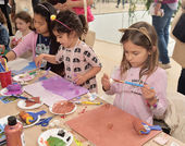 Several children stand at work at a table with art supplies, paint, and paintbrushes.