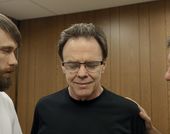 Three light-skinned people with short brown hair are seen from chest up, wearing t-shirts. The person on the right is seen in side view and places a hand on the shoulder of the person in the middle, who faces the camera, wears glasses and looks downward with a slight smile. The person on the left is visible from the side, has a dark beard, and looks across to the person on the right.