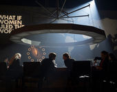 A circle of women around a table with a large circular light fixture above, and the words What if Women Ruled the World on the wall