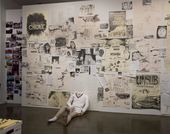 A gallery wall covered in drawings, with a headless mannequin sitting against it