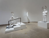 Life-sized white plaster figures of a girl climbing the stairs and a principal with arms crossed, both with antlers on their heads 