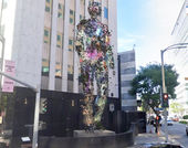 Rendering of a towering digital sculpture of a figure standing on an outdoor plinth