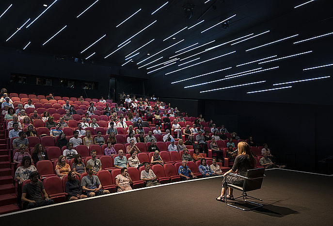 An audience sitting in a darkened theater, watching a woman on stage